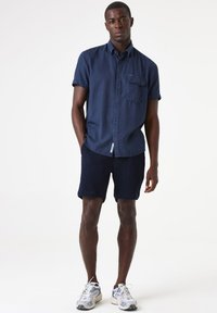Chemise boutonnée à manches courtes bleu marine avec une poche avant, associée à un short texturé bleu marine et des chaussures de sport blanches avec des accents bleus.