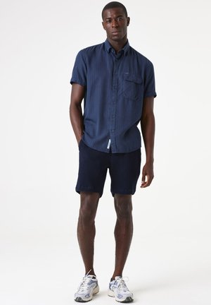 Camisa de manga corta azul marino con botones y un bolsillo frontal, combinada con pantalones cortos azul marino texturizados y zapatillas deportivas blancas con detalles azules.