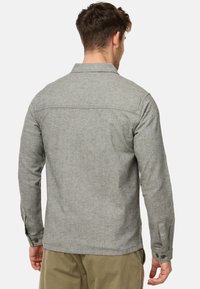 Homme portant une chemise à manches longues gris clair et un pantalon kaki, vu de dos sur un fond uni.