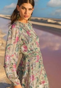 Rochie florală cu talie strânsă, având o bază verde deschis, accente roz și metalice argintii, și mâneci lungi, vaporoase.