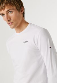 Giovane uomo con capelli castano chiaro che indossa una maglia bianca a maniche lunghe con un piccolo logo "Pepe Jeans London" sul petto e una piccola toppa sulla manica.