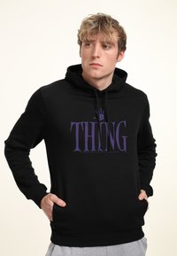 Giovane con i capelli biondi che indossa un felpa nera con la scritta "THING" e un impronta di mano viola, con le mani nelle tasche, che guarda di lato.