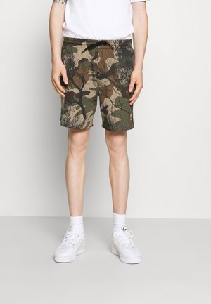 Person trägt grüne und braune Camouflage-Shorts, weiße Crew-Socken, weiße Adidas-Sneaker und ein weißes T-Shirt vor einem einfarbigen Hintergrund.