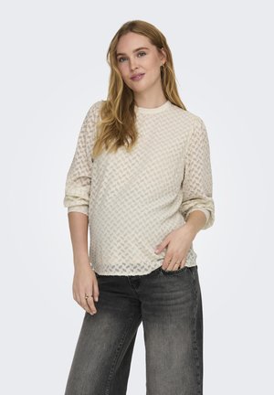 Cream-colored textured top with geometric patterns, round neckline, and three-quarter sleeves, paired with dark gray wide-leg jeans.
