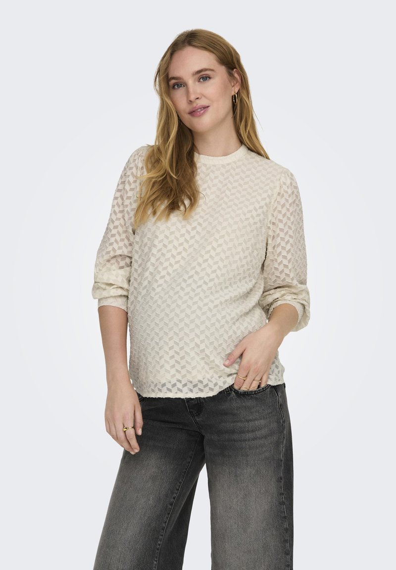 Top di colore crema con texture e motivi geometrici, scollatura rotonda e maniche a tre quarti, abbinato a jeans a gamba larga di un grigio scuro.
