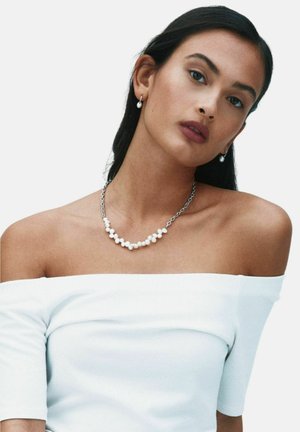 Moteris tamsiais plaukais, dėvinti perlinius auskarus lašo formos ir sidabrinę grandinėlę su sukrautomis baltomis perlėmis, dėvinti baltą palaidinę be pečių.