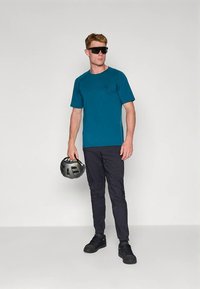 Türkises Kurzarm-T-Shirt mit rundem Ausschnitt, schwarze Hose mit Taschen, schwarze Schuhe und ein schwarzer Fahrradhelm, der in der linken Hand gehalten wird.