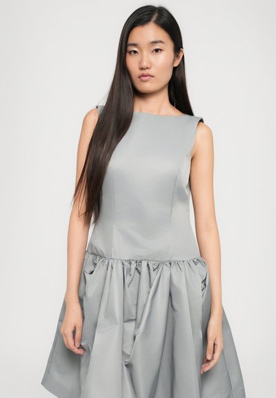 Szürke ruha, testhez simuló felső résszel és bő, rakott szoknyával. Simaságot biztosító anyagból készült. Ujjatlan fazon, kerek nyakkivágással és diszkrét fényléssel.