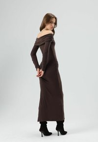 Bruine off-shoulder jurk met lange mouwen, gerimpelde details en een nauwsluitend ontwerp, gecombineerd met zwarte hoge laarzen met spitse neus.