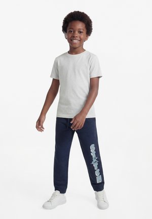 Gutt som smiler, iført lys grå kortermet skjorte, marineblå treningbukser med teksten "Stitch" og figurgrafikk, og hvite joggesko, stående.