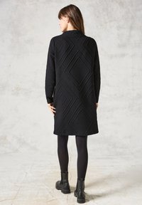 Zwarte gequiltte jurk met een gestructureerd ruitpatroon, lange mouwen en een ronde kraag, gecombineerd met zwarte enkellaarsjes en maillot.