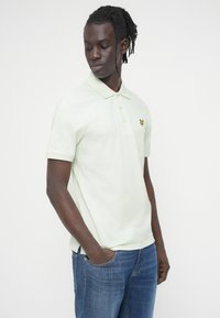 Ανοιχτό πράσινο πόλο μπλουζάκι με κοντά μανίκια, με κίτρινο λογότυπο πεταλούδας στο αριστερό στήθος, σε συνδυασμό με μπλε τζιν παντελόνι.