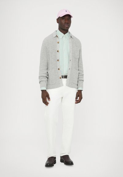 Pilkas kardiganas su mediniais mygtukais ant šviesiai žaliai dryžuotos palaidinės, kartu dėvimi balti kelnės ir tamsiai rudi batai. Ant galvos rožinė kepurė.