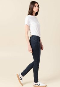 Jeans ajustados de denim azul oscuro, con una cintura alta, costuras mínimas y bolsillos frontales, combinados con una camiseta blanca lisa y zapatillas.