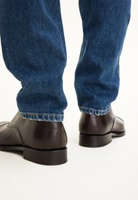Braune Lederschuhe mit glatter Oberfläche, getragen mit dunkelblauen Jeans, die am Saum leicht ausgefranst sind.