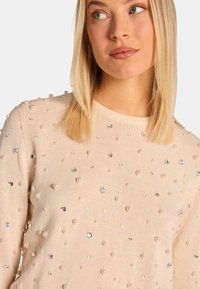 Beige gestrickter Pullover mit strukturiertem Oberflächen und verstreuten klaren und perlenartigen Verzierungen. Rundhalsausschnitt und lange Ärmel.