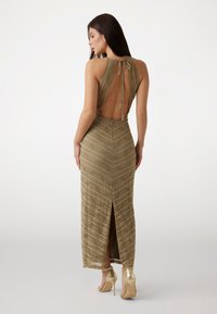 Abito maxi in maglia di un verde oliva metallico, con una profonda scollatura sulla schiena, dettaglio con nodo e motivo a zigzag con spacco alto. Abbinato a tacchi metallici.