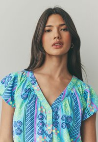 Chemise à manches courtes, col en V, de couleur bleue avec des motifs tourbillonnants et géométriques colorés ; elle possède un devant boutonné et des détails froncés aux épaules.