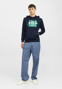 Donkerblauwe hoodie met een voorzak, voorzien van een groen grafisch logo in het midden. Gedragen met lichtblauwe jeans en witte sneakers.