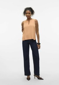 Top senza maniche color pesca con dettaglio a chiave, abbinato a jeans svasati blu scuro. La modella indossa décolleté marroni a punta e un bracciale nero.