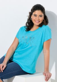Camiseta de manga corta en color turquesa con un dibujo lineal de elefantes en el frente, combinada con jeans oscuros, sentada sobre una superficie blanca.