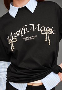 Černé tričko s nápisem "Mostly Magic", ozdobené perlovými akcenty tvořícími mašle. Přes něj je vrstveno modré pruhované košilové tričko.