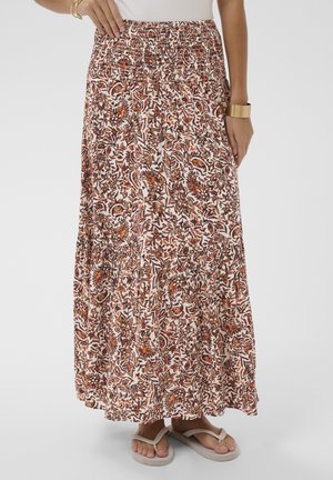 Žena má na sebe dlhú, voľne padajúcu sukňu s oranžovým a hnedým kvetinovým vzorom, v kombinácii s béžovými sandálmi a zlatými náramkami.