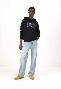 Felpa nera con la scritta "JACK & JONES", abbinata a jeans baggy blu chiaro e scarpe gialle. Design semplice, stile casual, texture morbida.