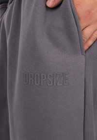Grå joggingbukser lavet af blødt stof med ribbede detaljer og et synligt præget "DROPSIZE" logo på venstre sidelomme.
