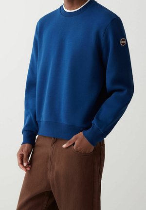 Mężczyzna w niebieskiej bluzie z okrągłym dekoltem z małą naszywką z logo na górnej części rękawa oraz w brązowych spodniach, z ręką włożoną do kieszeni.