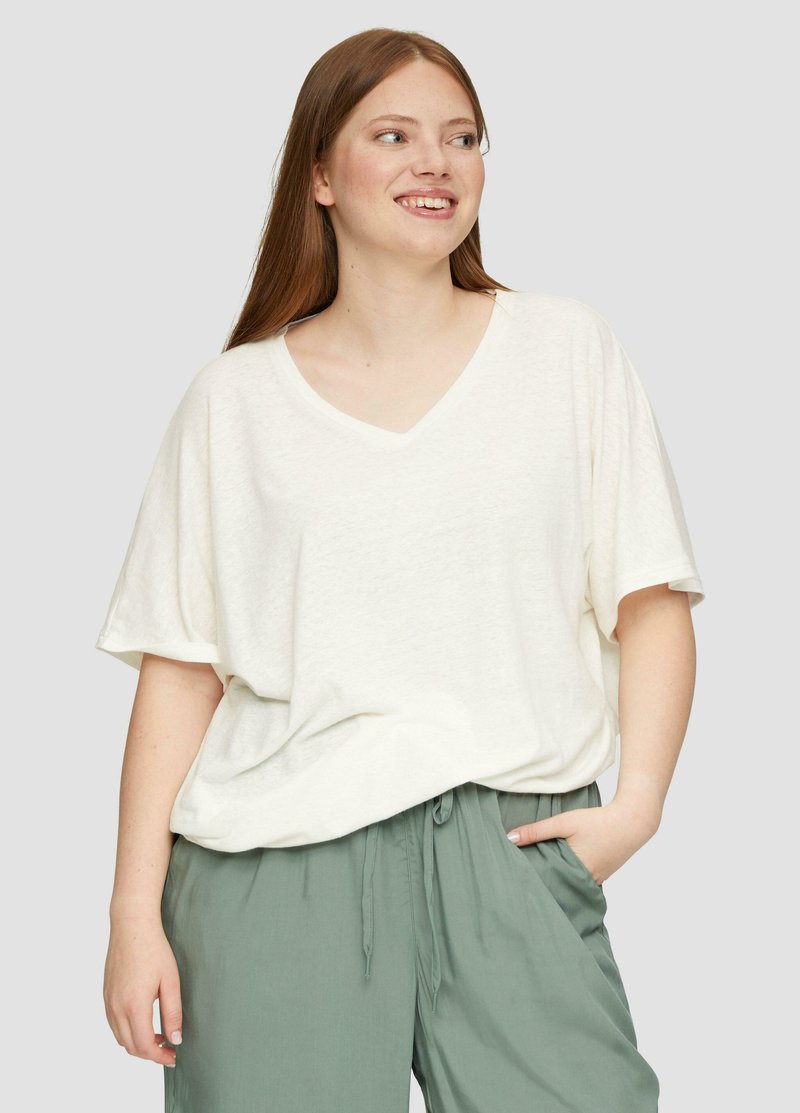 Witte, losse, korte mouw V-hals shirt van zachte stof, gecombineerd met lichtgroene broek met trekkoord, op een neutrale achtergrond.