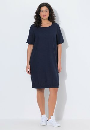 Vrouw staand, gekleed in een losse marineblauwe jurk tot op de knie met korte mouwen en witte sneakers, tegen een effen lichte achtergrond.