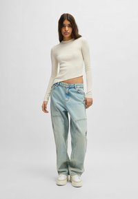 Top a maniche lunghe a coste di colore crema abbinato a jeans baggy blu chiaro. I jeans presentano tasche laterali e un leggero effetto acid-wash. Sneakers bianche.