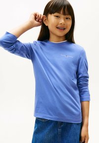 Junges Mädchen mit langen dunklen Haaren, das ein blaues Tommy Hilfiger Langarmshirt und einen Jeansrock trägt, lächelt mit Zahnspange und hält die Hand nah am Ohr.