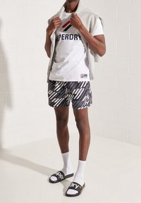 Bílá tričko s logem "Superdry", šedý svetr položený na ramenou, vzorované plavkové kraťasy v černobílém provedení, bílé ponožky, černé sandály.