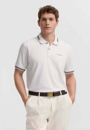 Uomo che indossa una polo bianca a maniche corte con colletto e polsini a righe nere, pantaloni bianchi e cintura scura con fibbia in ottone, mani in tasca.