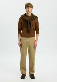 Jeune homme portant un pull marron avec une écharpe marron foncé drapée sur les épaules, un pantalon beige et des chaussures marron foncé, debout devant un fond blanc.