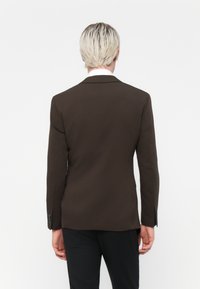 Sacou tailored maro, cu o deschidere simplă, rever cu crestătură și închidere cu două butoane, prezentat din spate pe un model care poartă pantaloni negri.
