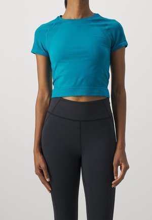Persona que lleva una camiseta deportiva corta de manga corta en turquesa y mallas negras de cintura alta, de pie con los brazos relajados a los lados.