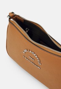 Borsa a tracolla marrone con texture, dotata di chiusura a zip, caratterizzata da un logo metallico e dettagli di cucitura lungo la parte superiore. L'interno è nero.
