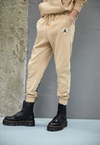 Beige Jogginghose mit einem Jordan-Logo und bedrucktem Text am Bein, kombiniert mit schwarzen Stiefeln mit dicker Sohle und Schnürdetails.