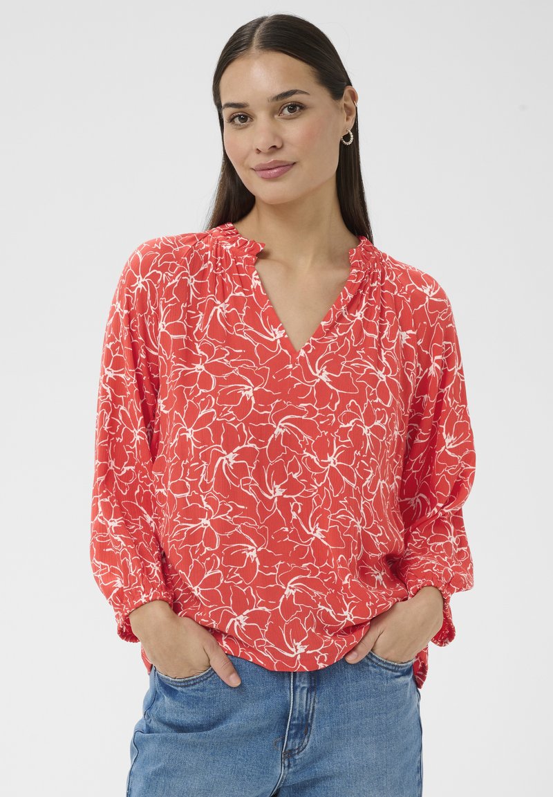 Femme aux longs cheveux noirs portant un chemisier rouge à fleurs avec col en V et un jean bleu, mains dans les poches, fond gris neutre.