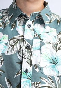 Camicia a maniche corte a bottoni floreale in teal, con motivi di ibisco bianchi e foglie verdi, dotata di taschino sul petto e bottoni grigi. Tessuto in cotone.