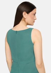 Blouse sans manches verte avec une texture lisse, un col arrondi et une fermeture éclair discrète au dos.