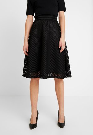 Femme portant une jupe noire texturée mi-longue en ligne A, un haut noir à manches courtes et des escarpins noirs pointus, debout sur un sol blanc.