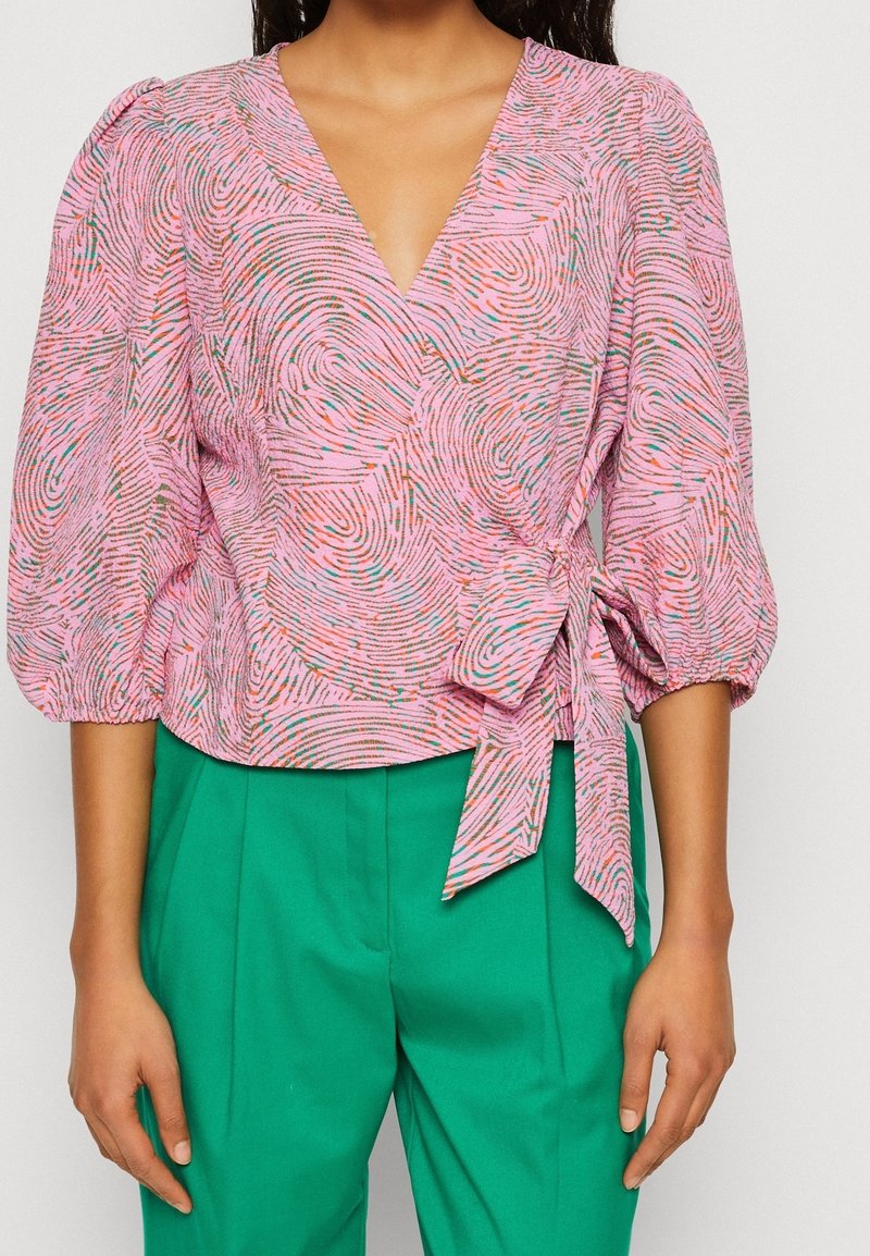 Femme portant un chemisier cache-cœur rose avec un motif tourbillonnant multicolore et des manches bouffantes, associé à un pantalon vert taille haute.
