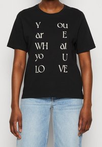 Camiseta de algodón negra con mangas cortas que presenta texto blanco dispuesto verticalmente, escribiendo "ERES QUIEN AMAS" en varios estilos.