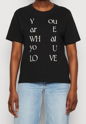 Camiseta de algodón negra con mangas cortas que presenta texto blanco dispuesto verticalmente, escribiendo "ERES QUIEN AMAS" en varios estilos.