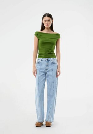 Ung kvinde iført en grøn off-shoulder top, lyseblå vide bukser og brune platformsko, stående mod en hvid baggrund.