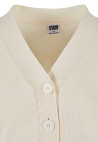 Beżowy sweter dzianinowy z dekoltem w serek, dwoma białymi guzikami i metką "Urban Classics", rozmiar XS, wyprodukowano w Bangladeszu.